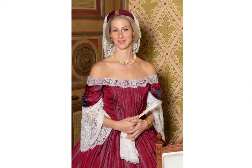 Die bestehende Beschreibung lautet: "Das Bild zeigt die Stadtführerin." Hier ist die optimierte Version: "Stadtführerin in historischer Tracht vor kunstvollem, antikem Hintergrund stehend."
