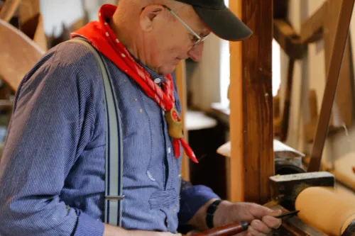 Symbolbild Drechsler, älterer Mann mit schwarzer Schildkappe, blau-weisses Hemd, rotes Halstuch. Er hat ein Stecheisen in der Hand und steht an einer alten Drechslermaschine