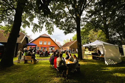 Menschen sitzen an Tischen und Bänken im Garten vor einem roten Backsteinhaus unter Bäumen bei einer Veranstaltung mit Bühne und Pavillons