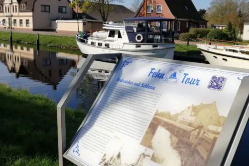 Infotafel zum historischen Rundgang vor einem Kanal mit Boot und Häusern in Rhauderfehn