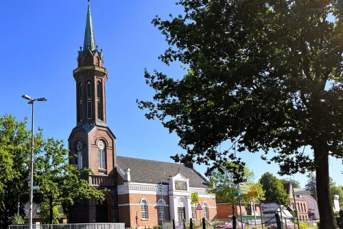 Kirchturm mit spitzem Dach neben Bäumen und einem Gebäude mit weißer Fassade unter blauem Himmel
