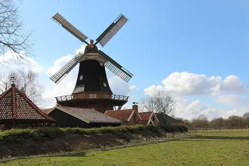 Windmühle steht auf einer Wiese unter bewölktem Himmel mit tiefstehender Sonne, links Bäume mit kahlen Ästen.