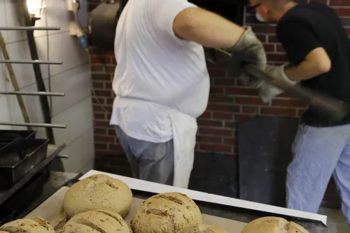 Symbol Backtag, im Vordergrund liegen mehrere runde gebackene Brotlaibe, im Hintergrund steht ein älterer Mann mit einer weissen Backschürtze. Dieser hat einen langen Brotschieber aus Holz in der Hand. Vor ihm steht ein weiterer Mann. Beide stehen vor einem Steinbackofen.