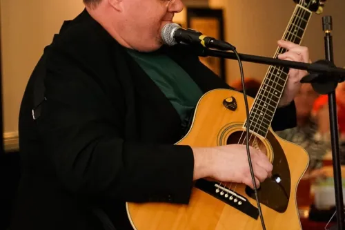 Person sitzt und spielt akustische Gitarre vor Mikrofon in Innenraum mit unscharfem Hintergrund