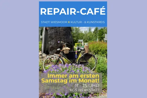 Das Plakat zeigt ein schwarzes Fahrrad steht vor lila Blüten, im Hintergrund grüner Wald. Im oberen Bereich des Plakates in einem blauen Kasten steht Repair-Cafe, Stadt Wiesmoor eine gelbes X dann steht da weiter Kultur- und Kunstkreis. In unteren Bereich des Plakates steht in einem sehr hellen grauen Kasten "Immer an dem ersten Samstag im Monat. 11-15 Uhr in der KGS Werkstatt.