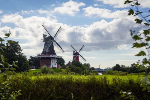 Zwei historische Windmühlen in Greetsiel, umgeben von Wiesen, Bäumen und weitem Himmel.