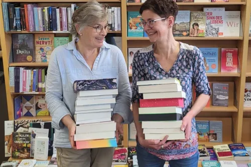 Zwei Frauen stehen vor Bücherregalen mit den Überschriften 'Das besondere Buch' und 'Bestseller' und halten jeweils einen großen Stapel Bücher.