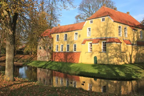 Gelbes Backsteingebäude mit rotem Dach, umgeben von Wassergraben und herbstlich belaubten Bäumen.