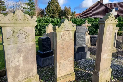 Drei verwitterte Sandstein-Gedenksteine mit Reliefs und Inschriften auf einem Friedhof, im Hintergrund Hecke und Haus mit rotem Ziegeldach.