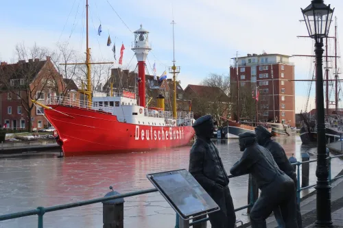 Rotes Feuerschiff mit der Aufschrift »Deutsche Bucht« im Hafen, davor zwei bronzene Figuren an einem Geländer und eine Laterne.