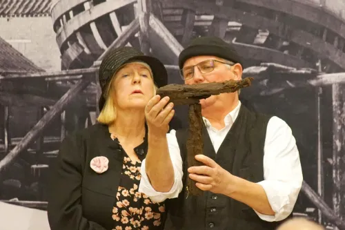 Zwei Personen vor einem historischen Hintergrund halten ein großes, rostiges Kreuz aus Metall