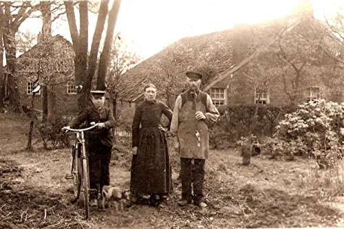 Drei Personen in historischer Kleidung stehen vor einem Reetdachhaus, eine Person hält ein Fahrrad, eine kleine Kuh ist zu ihren Füßen.