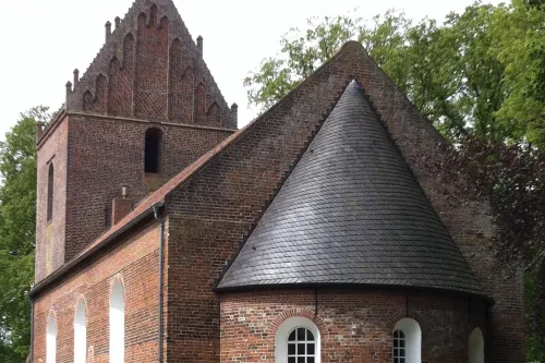Backsteinkirche mit romanischem Rundturm und gotischem Giebel, umgeben von Bäumen