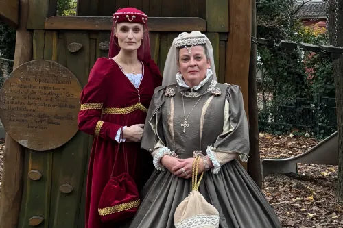 Zwei Frauen in historischen Kleidern posieren im Freien vor einer hölzernen Kulisse im Herbst.
