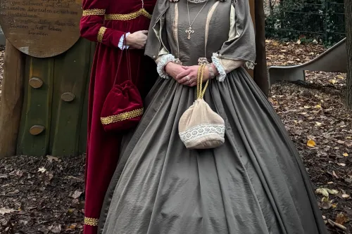 Zwei Frauen in historischen Kleidern posieren im Freien vor einer hölzernen Kulisse im Herbst.