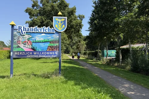 Willkommensschild mit dem Schriftzug 'Rhauderfehn' und 'Herzlich willkommen zwischen Moor und Meer' neben einem Weg mit zwei Radfahrern und Bäumen im Hintergrund