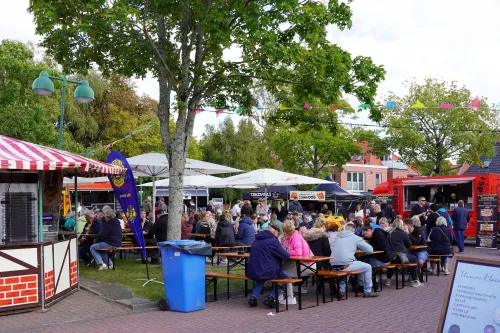 Menschen sitzen an Holzbänken zwischen Foodtrucks und bunten Fahnen beim Street Food Festival.