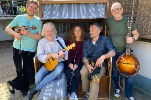 Fünf Personen mit Musikinstrumenten sitzen und stehen um eine Strandkorbbank auf gepflastertem Boden.