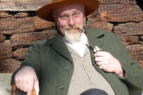 Mann in historischer Kleidung mit Pfeife und Stock sitzt lächelnd vor einem Holzhaus in der Sonne.