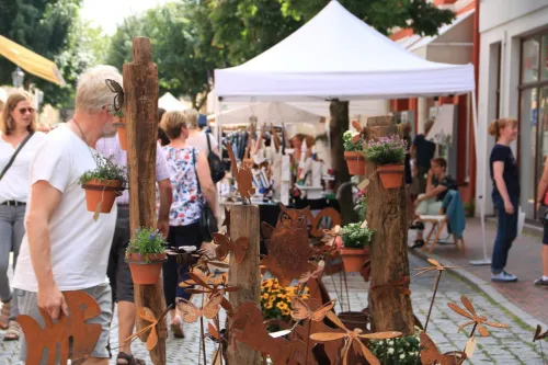 Belebter Kunst- und Handwerkermarkt mit Besuchern, weißen Ständen und rostiger Gartendeko.