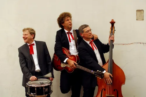 Drei Musiker im Anzug mit roter Fliege posieren mit Trommel, Gitarre und Kontrabass.