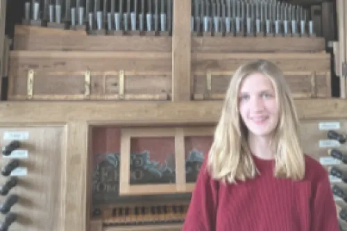 Junge Frau in rotem Pullover lächelt vor der großen, hölzernen Pfeifenorgel in der Pilsumer Kirche.