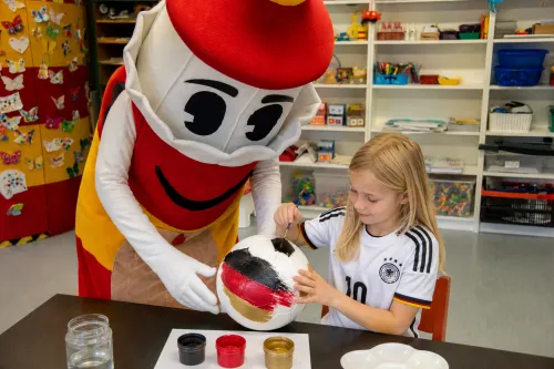 Ein Mädchen im Deutschland-Trikot bemalt mit dem Maskottchen Lükko einen Fußball in Schwarz-Rot-Gold.