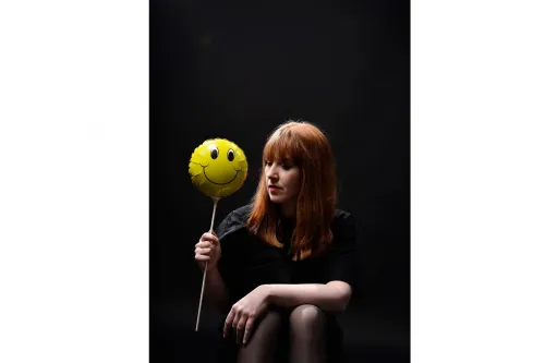 Frau mit rotem Haar hält gelben Smiley-Ballon vor dunklem Hintergrund.