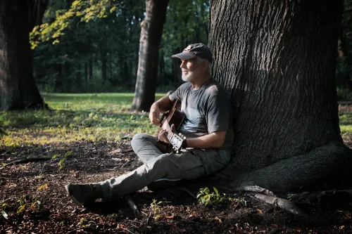 Gitarrist sitzt im Wald an einem Baumstamm und spielt im Sonnenlicht.
