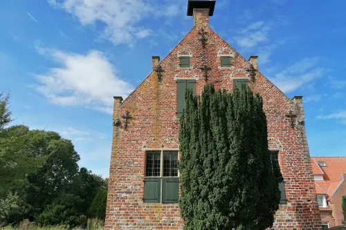 Backsteinbau mit grünen Fensterläden und markanter Giebelfront, teils verdeckt durch hohen Baum.