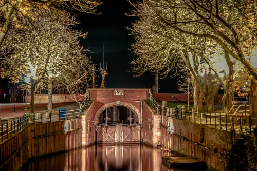 Beleuchtete Schleuse in Greetsiel, umgeben von kahlen Bäumen, spiegelt sich im ruhigen Wasser der Nacht.