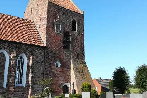 Backsteinkirche mit hohem Turm und rotem Ziegeldach vor blauem Himmel, umgeben von Grabsteinen.
