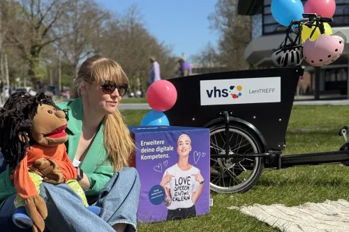 Eine Frau mit blondem Pferdeschwanz, Sonnenbrille, grünem Blazer und Jeans sitzt auf einer Wiese und hält eine bunte Handpuppe mit schwarzen Zöpfen. Neben ihr steht ein Sitzwürfel mit der Aufschrift „Erweitere deine digitale Kompetenz“ und einem Bild einer jungen Person, die ein T-Shirt mit der Aufschrift „Love Speech Entwicklerin“ trägt. Im Hintergrund steht ein schwarzes Lastenrad mit Luftballons und dem Logo „vhs LernTREFF“. Das Wetter ist sonnig, Bäume ohne Laub und ein modernes Gebäude sind im Hintergr