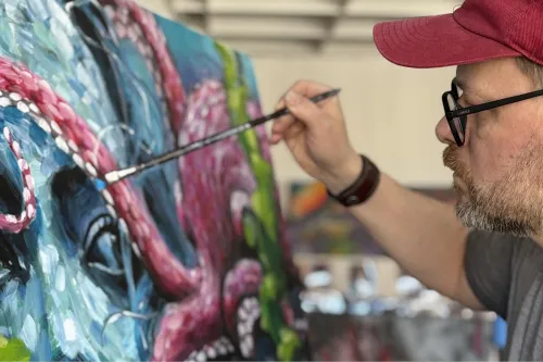 Mann malt auf einer Leinwand einen Octopus im Wasser, während man in diesem ein Frauengesicht erkennen kann