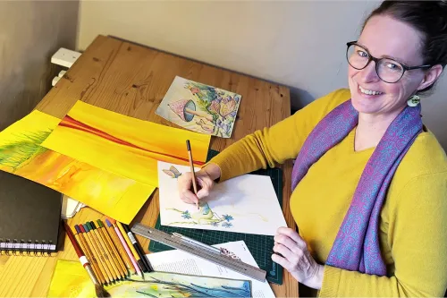 Frau mit gelben Pullover sitzt am Schreibtisch und schaut in die Kamera. Auf dem Schreibtisch liegen verschiedene gemalte Bilder.