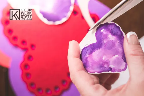 ein getuschtes, lila Herz aus Papier wird mit einer Schere ausgeschnitten