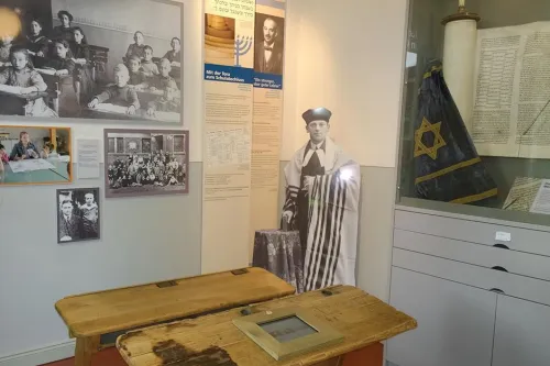 Im Vordergrund stehen zwei historische Schulbänke. Im Hintergrund ist eine Torarolle zu sehen sowie Fotos aus historischen Klassenzimmern und ein Fotoaufsteller des vorletzten Lehrers der Gemeinde August-Gottschalk