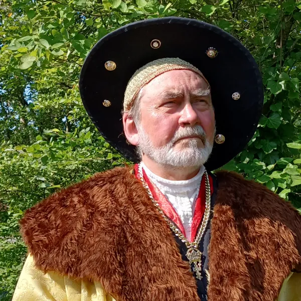 Mann in historischer Kleidung mit Pelzkragen und großem Hut vor grünem Hintergrund im Freien.