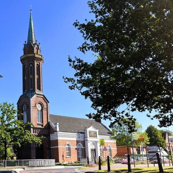 Kirchturm mit spitzem Dach neben Bäumen und einem Gebäude mit weißer Fassade unter blauem Himmel