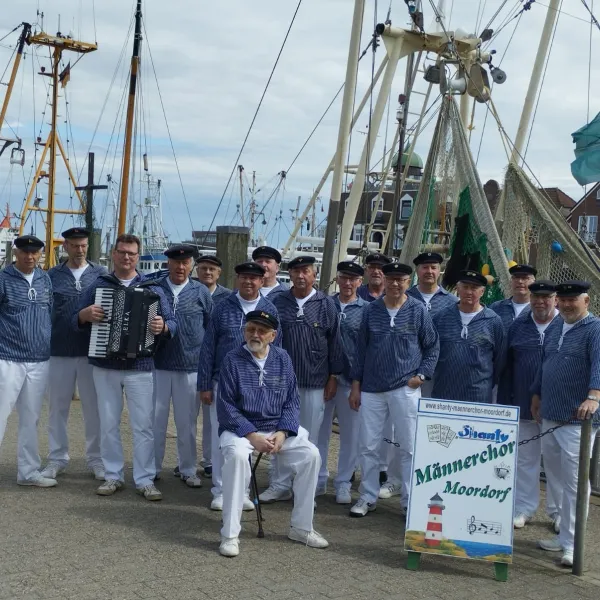 eine Gruppe von 17 Männern in blauen Seemannshemden und weißer Hose mit Seemannsmütze. Im Hintergrund sieht man Bootsmasten.
