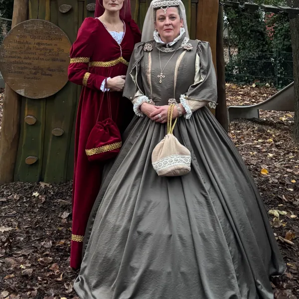 Zwei Frauen in historischen Kleidern posieren im Freien vor einer hölzernen Kulisse im Herbst.