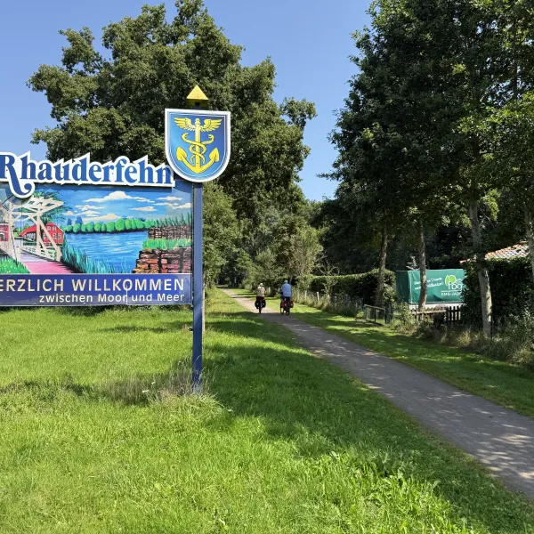 Willkommensschild mit dem Schriftzug 'Rhauderfehn' und 'Herzlich willkommen zwischen Moor und Meer' neben einem Weg mit zwei Radfahrern und Bäumen im Hintergrund