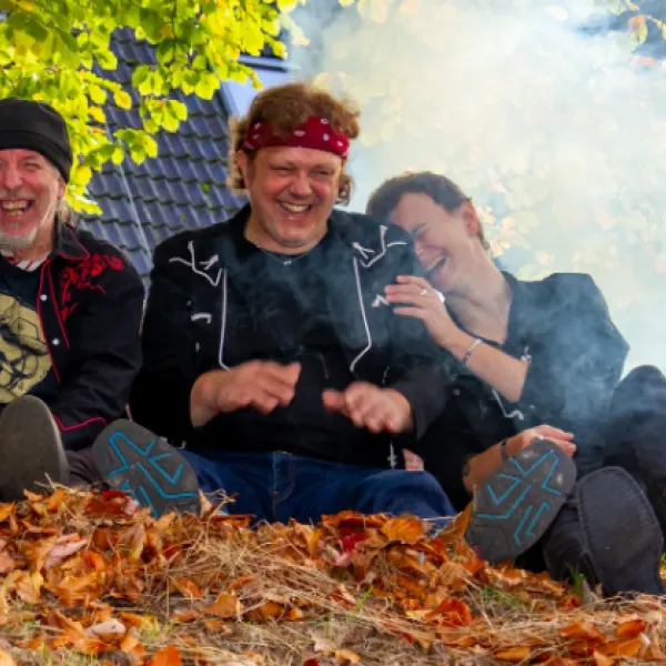 Drei Personen sitzen auf einem mit Herbstlaub bedeckten Boden vor einem Hausdach, umgeben von Rauch und Bäumen mit grünen und gelben Blättern