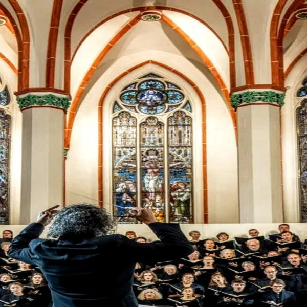 Ein Dirigent leitet einen großen Chor vor hellen Buntglasfenstern in einer Kirche.