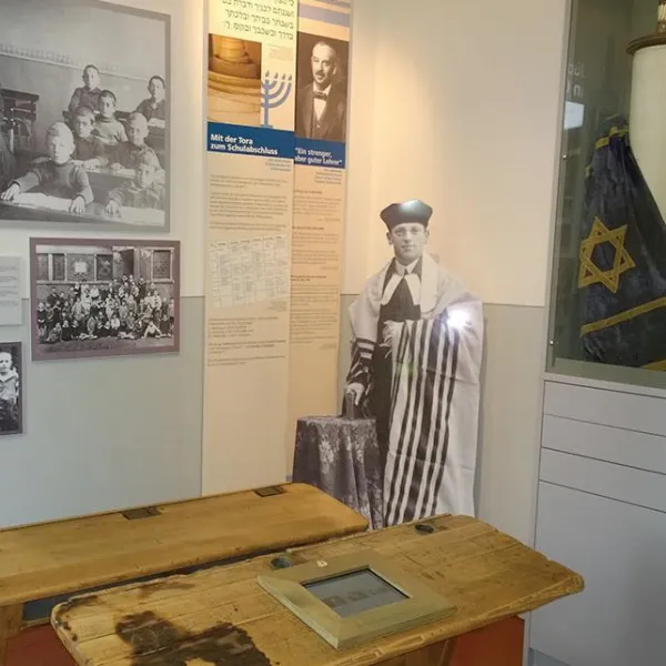 Im Vordergrund stehen zwei historische Schulbänke. Im Hintergrund ist eine Torarolle zu sehen sowie Fotos aus historischen Klassenzimmern und ein Fotoaufsteller des vorletzten Lehrers der Gemeinde August-Gottschalk