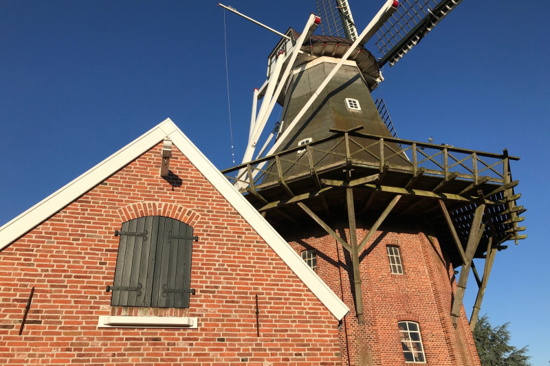 Historische Windmühle aus rotem Backstein mit Holzgalerie und weißen Flügeln unter blauem Himmel.