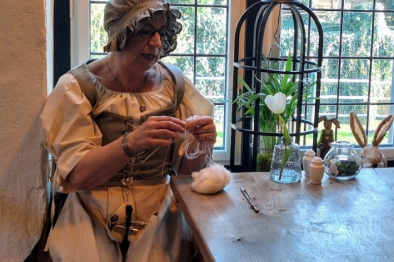 Frau in historischer Kleidung spinnt Wolle an Holztisch vor Fenster mit Frühlingsdekoration.
