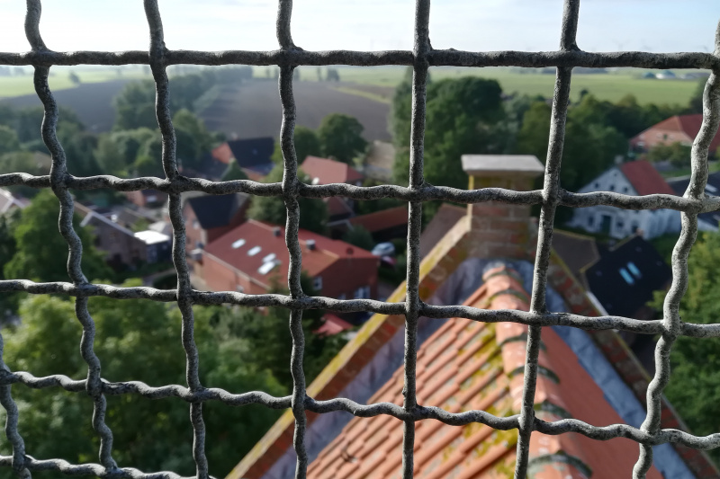 Blick durch das Schutzgitter eines Kirchturms auf rote Ziegeldächer und grüne Landschaft in Eilsum.