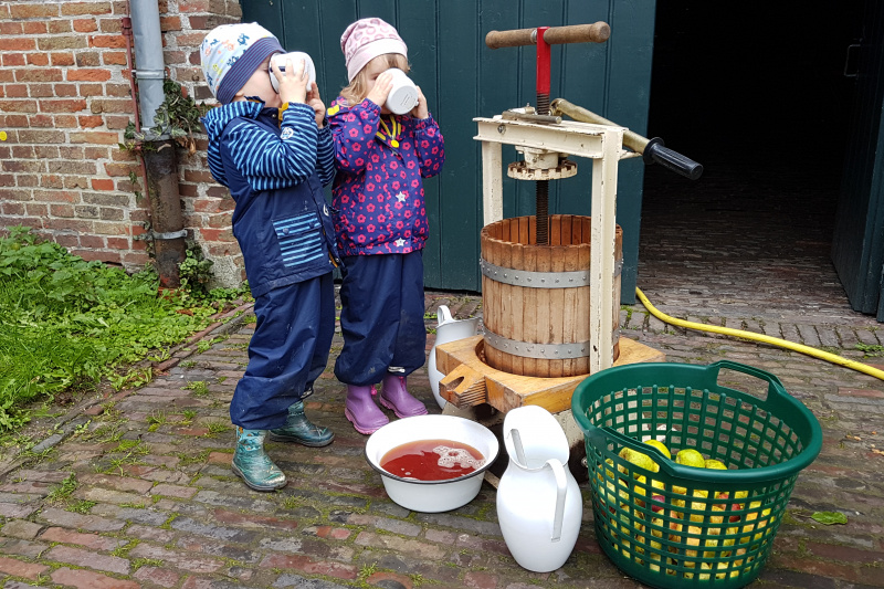 Zwei Kinder trinken frischen Apfelsaft neben einer alten Saftpresse beim Apfelfest in Campen.