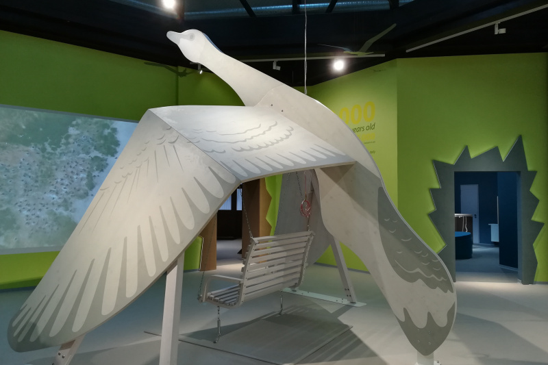 Große Holzskulptur einer fliegenden Gans mit Schaukel darunter in einer modernen Ausstellungshalle.